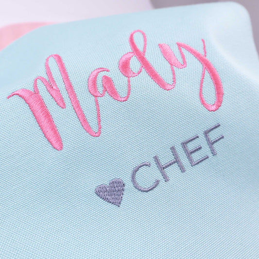 A close-up of a light blue fabric item with pink embroidered script reading 'Madly' and gray embroidered text reading 'CHEF' with a small gray heart.