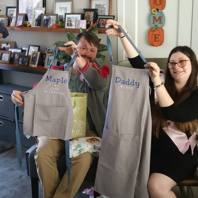 Two people holding aprons with personalized text in a home setting.