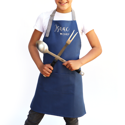 Child wearing a personalized navy apron over a white shirt, holding kitchen utensils