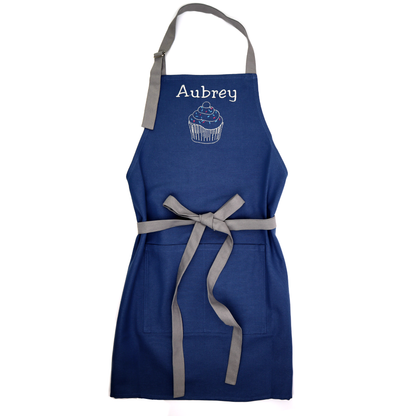 Personalized navy apron on a white background, tied bow, embroidered with the name "Aubrey" above a small icon