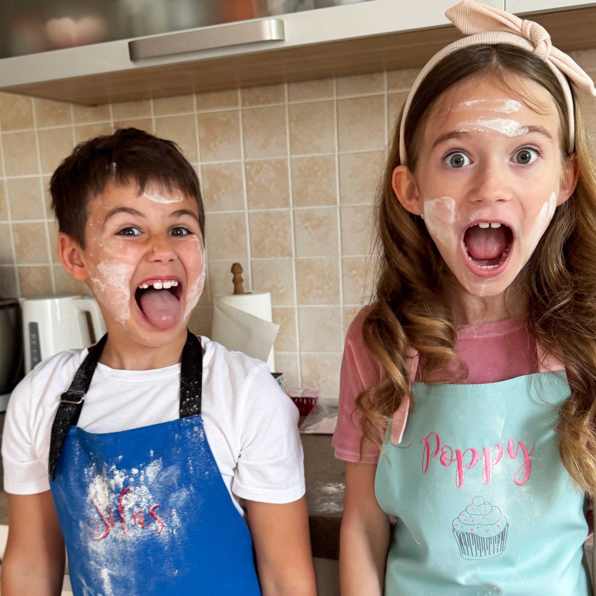 Two children wearing colorful baking aprons in a kitchen, covered in white flour and smiling with excited expressions. The boy wears a blue apron personalized with name Silas, and the girl wears a light blue apron with pink lettering that says 'Poppy' and a cupcake graphic.