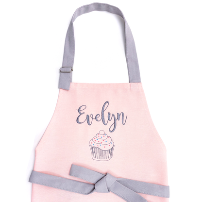 Personalized pink apron on a white background, embroidered with the name "Evelyn" above a cupcake design