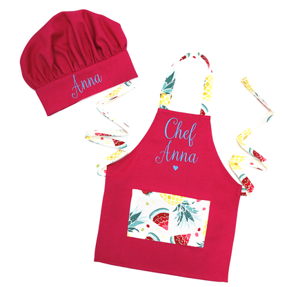 Personalized hot pink apron and chef hat set with a strawberry and pineapple print, embroidered "Chef Anna" in blue script, on a white background