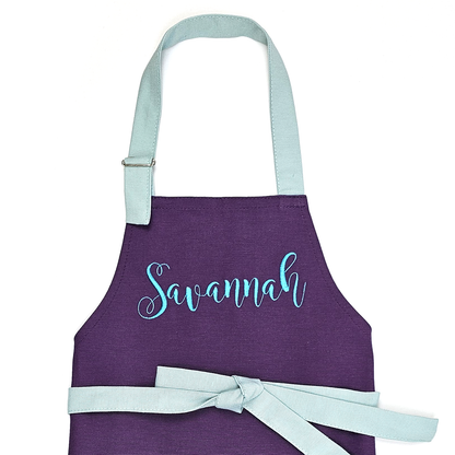 Personalized purple kids apron on a white background, embroidered with the name "Savannah" in turquoise cursive