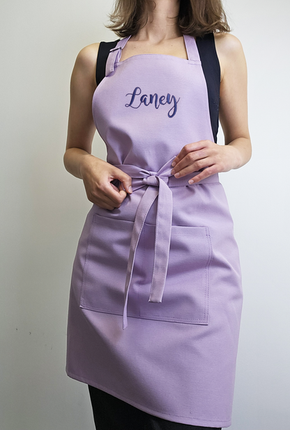 Adult woman wearing a personalized lavender apron, tying the front bow, embroidered with the name "Laney"