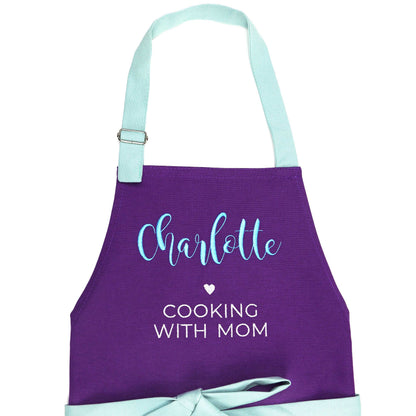 A purple kitchen apron with a light blue neck strap and waist tie, personalized with the name 'Charlotte' in light blue script and the phrase 'COOKING WITH MOM' in white text.