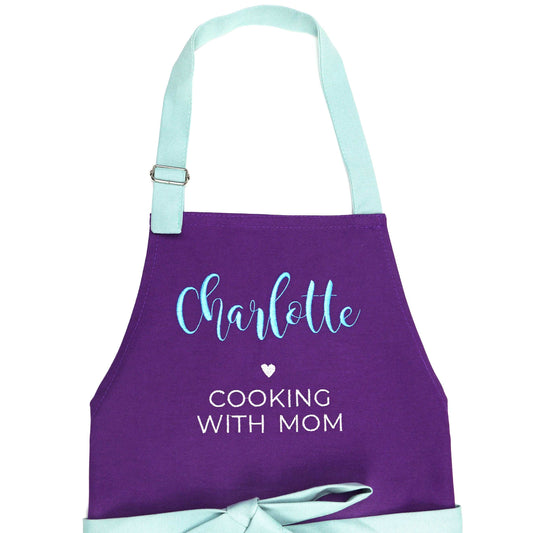 A purple kitchen apron with a light blue neck strap and waist tie, personalized with the name 'Charlotte' in light blue script and the phrase 'COOKING WITH MOM' in white text.