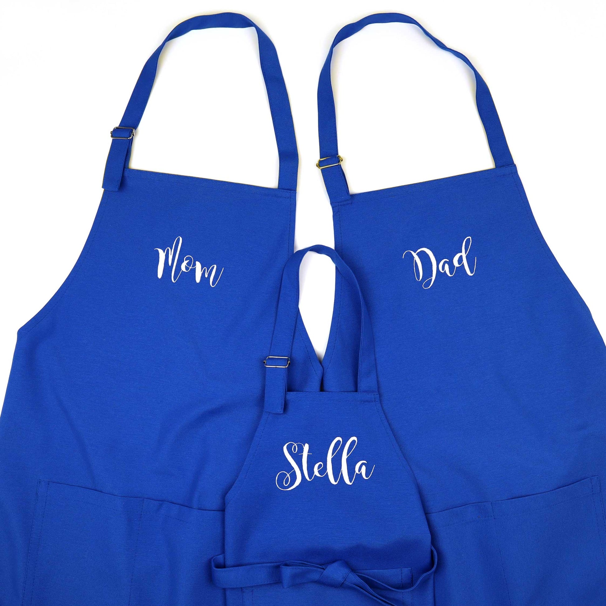 Three royal blue aprons with white script names 'Mom', 'Dad', and 'Stella' on a plain white background.