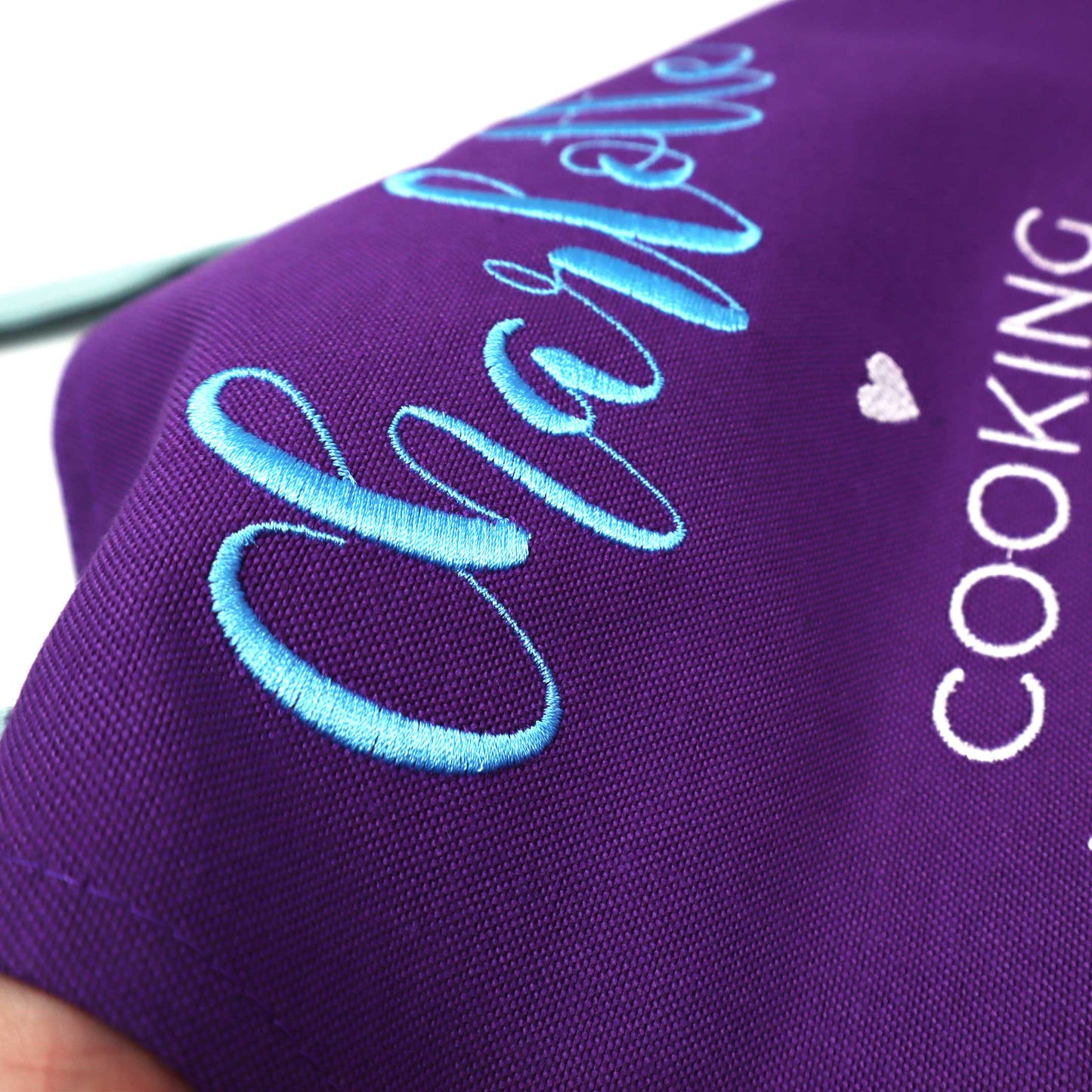 Close-up of a purple fabric item with light blue embroidered script reading 'Charlotte' and white text reading 'COOKING' alongside a small white heart.
