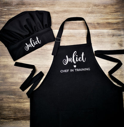 Personalized black apron and matching chef hat laid on a wooden surface, embroidered "Juliet ♥ Chef in Training"