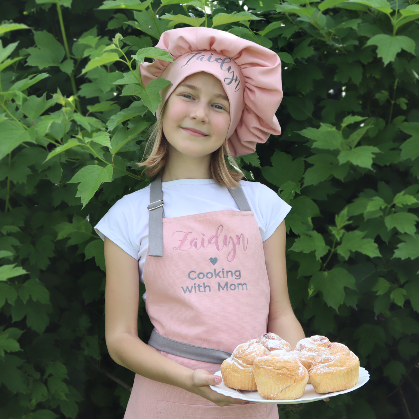 Child wearing a pink chef hat and apron holding a plate of pastries in front of green foliage.