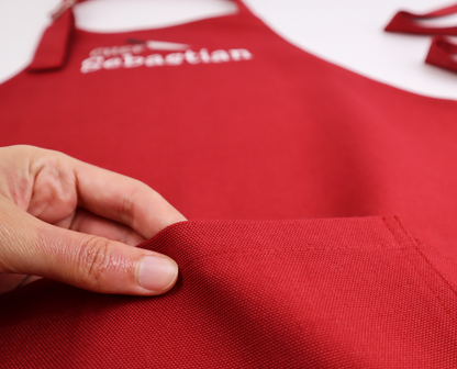 Close-up of a hand showing the fabric texture of a personalized dark red apron, embroidered with a name ending in "...bastian"
