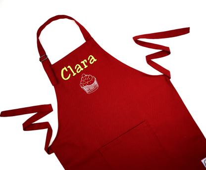 Personalized dark red apron with straps spread out, embroidered with the name "Clara" above a small icon