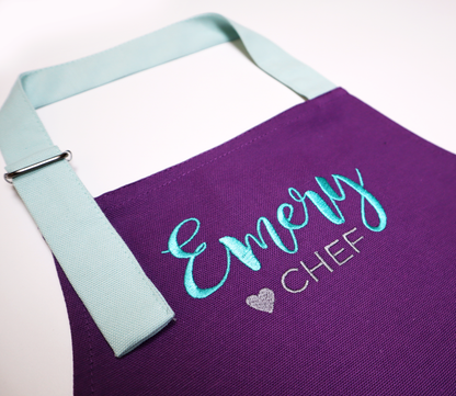 Close-up of "Emery ♥ Chef" embroidered on a personalized purple apron with a mint strap
