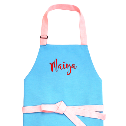 Personalized blue apron with a pink tied bow, embroidered with the name "Maiya" in red cursive