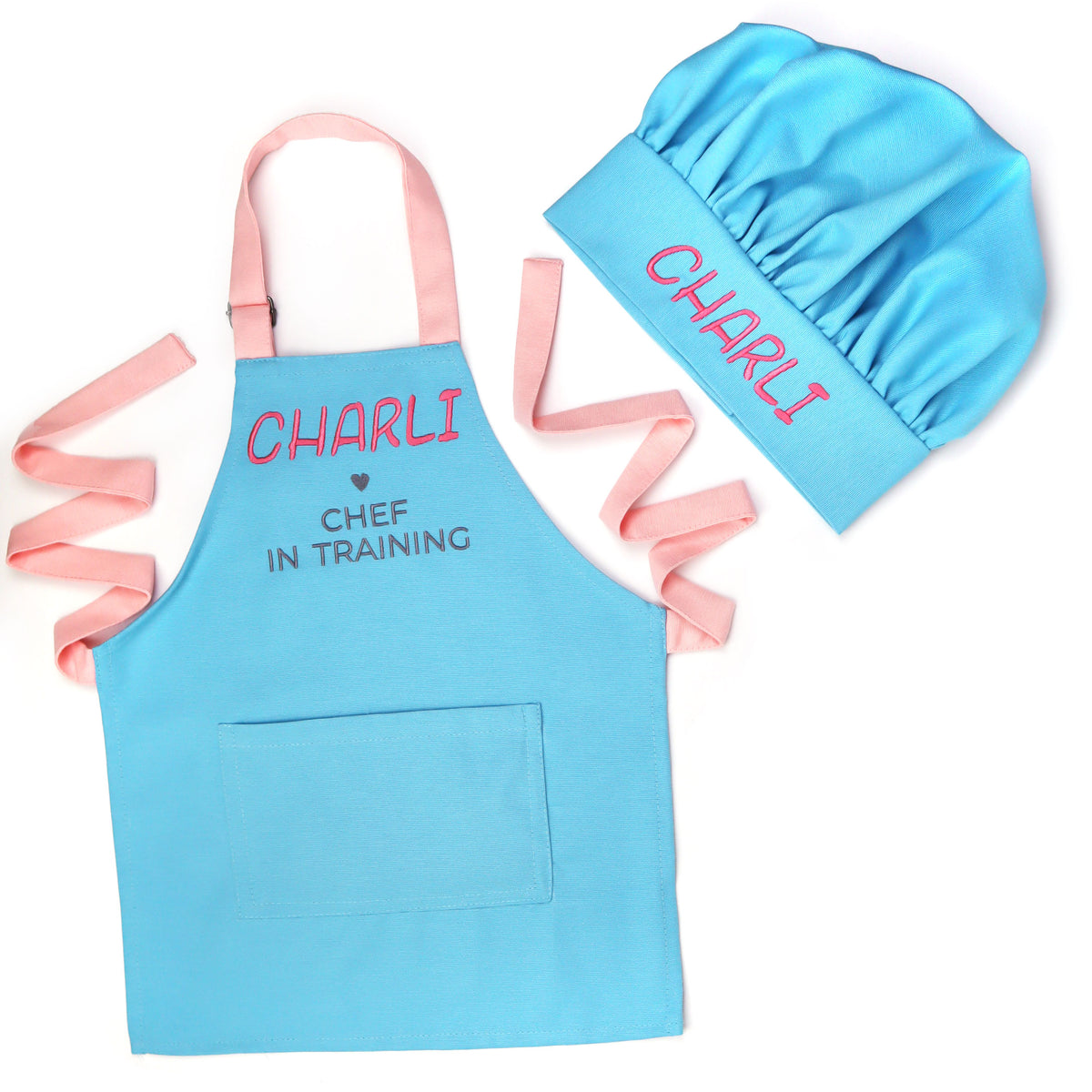 A children's cooking costume set featuring a light blue apron with pink neck and waist straps, and a matching light blue chef hat. The apron is printed with the text 'CHARLI' in pink and 'CHEF IN TRAINING' in gray.