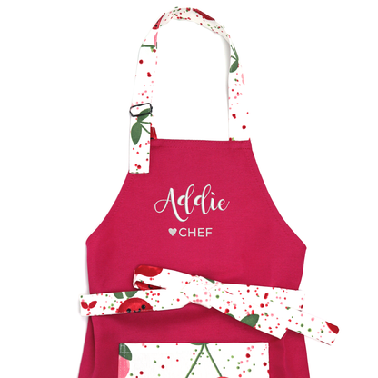 Personalized hot pink apron with cherry-print straps on a white background, embroidered "Addie ♥ CHEF"