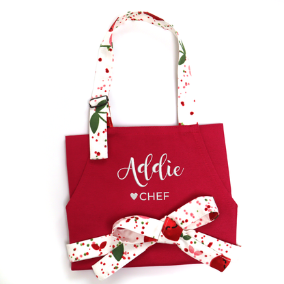 Personalized hot pink apron with a cherry-print bow tied at the front, embroidered "Addie ♥ CHEF", front pocket detail
