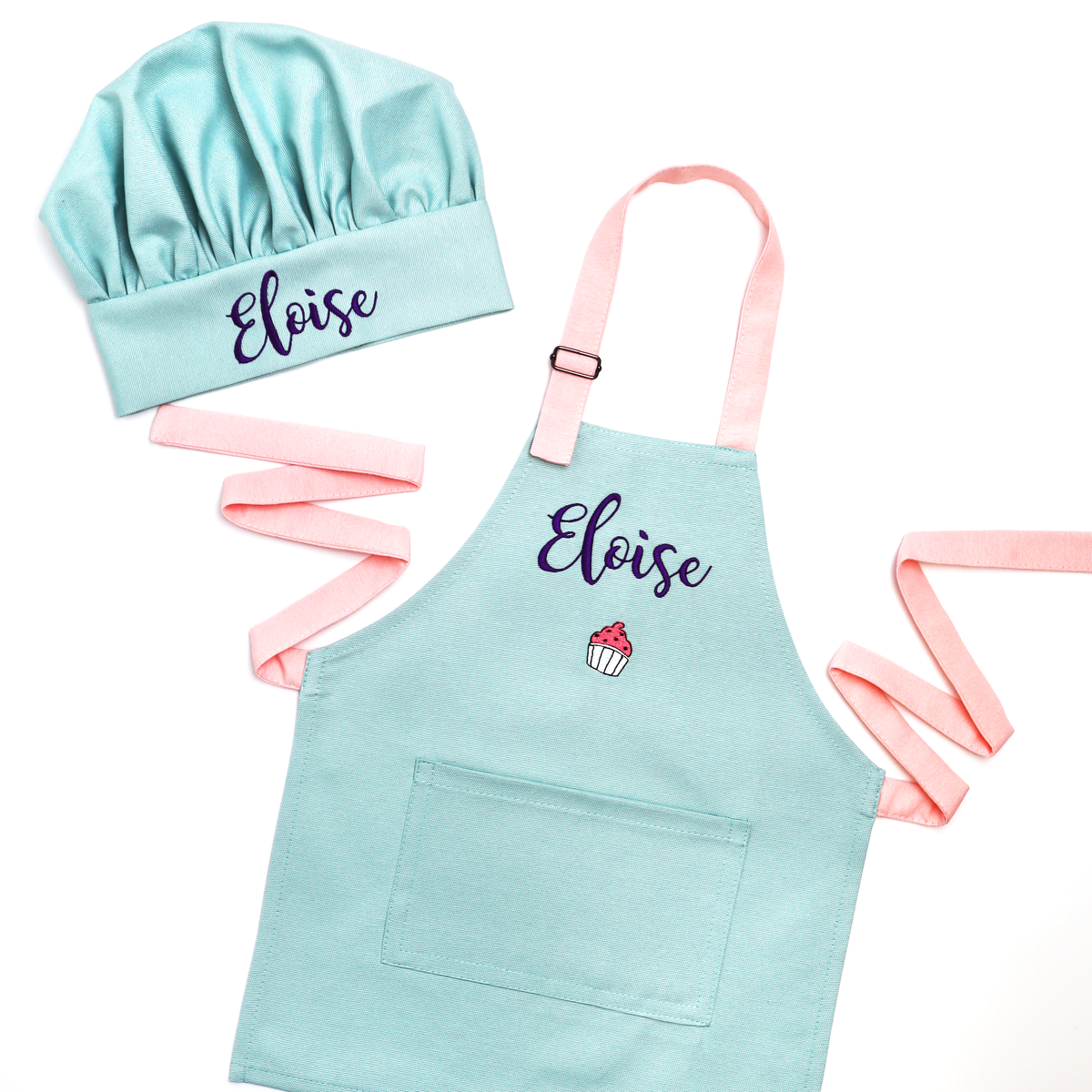 A children's chef hat and matching apron set in light mint with pastel pink straps, both personalized with the name 'Eloise' in dark purple script. The apron also features a small embroidered cupcake graphic.