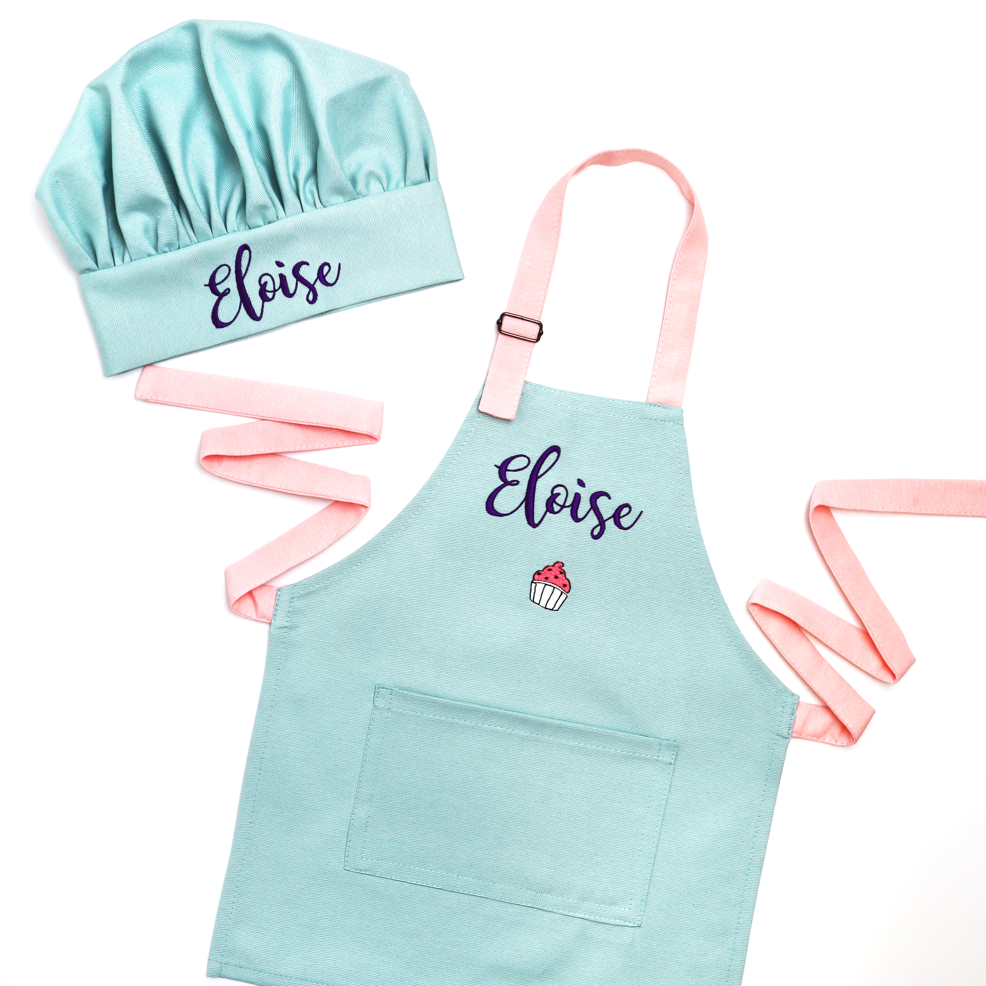 A children's chef hat and matching apron set in light mint with pastel pink straps, both personalized with the name 'Eloise' in dark purple script. The apron also features a small embroidered cupcake graphic.