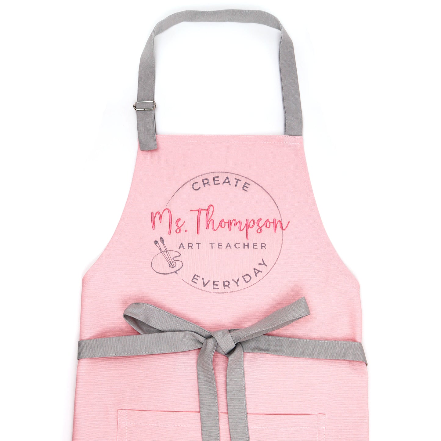 Pink personalized art teacher apron on dress form with grey adjustable straps and bow-tied waist, embroidered with coral script name Ms. Thompson Art Teacher in a circle stamp with painter palette and brushes