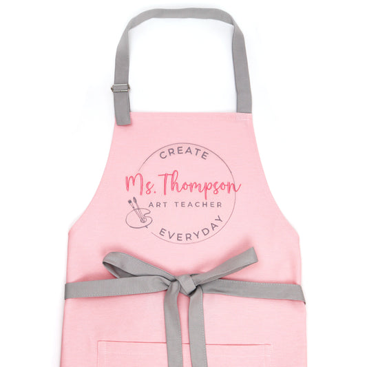 Pink personalized art teacher apron on dress form with grey adjustable straps and bow-tied waist, embroidered with coral script name Ms. Thompson Art Teacher in a circle stamp with painter palette and brushes