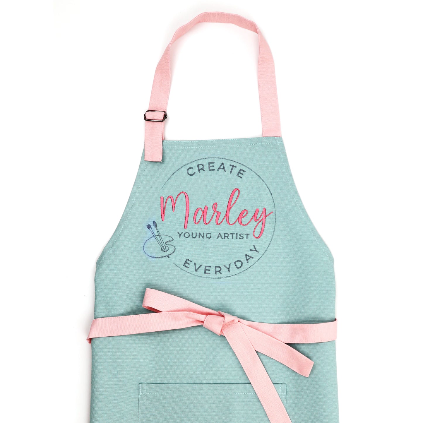 Mint green kids art apron with pink straps and embroidered "Create Everyday Young Artist" design with the name Marley in pink script and a small palette emblem, flat lay on white background
