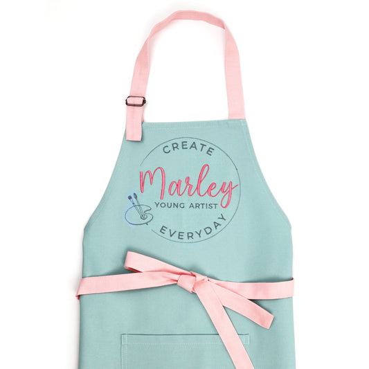 Mint green kids art apron with pink straps and embroidered "Create Everyday Young Artist" design with the name Marley in pink script and a small palette emblem, flat lay on white background