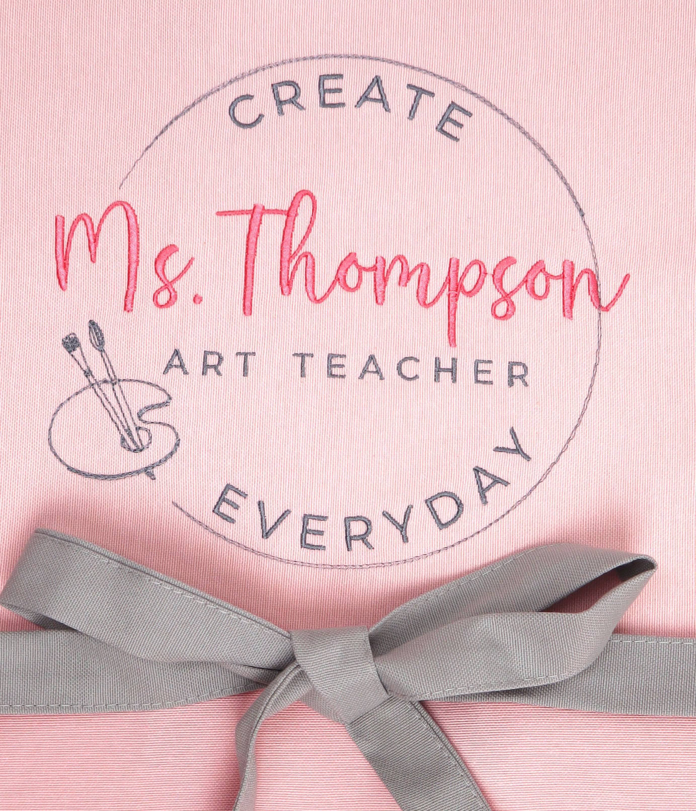 Close-up of embroidered art teacher apron showing coral script name Ms. Thompson with Art Teacher in grey thread, surrounded by Create Everyday circle stamp and embroidered painter palette with brushes