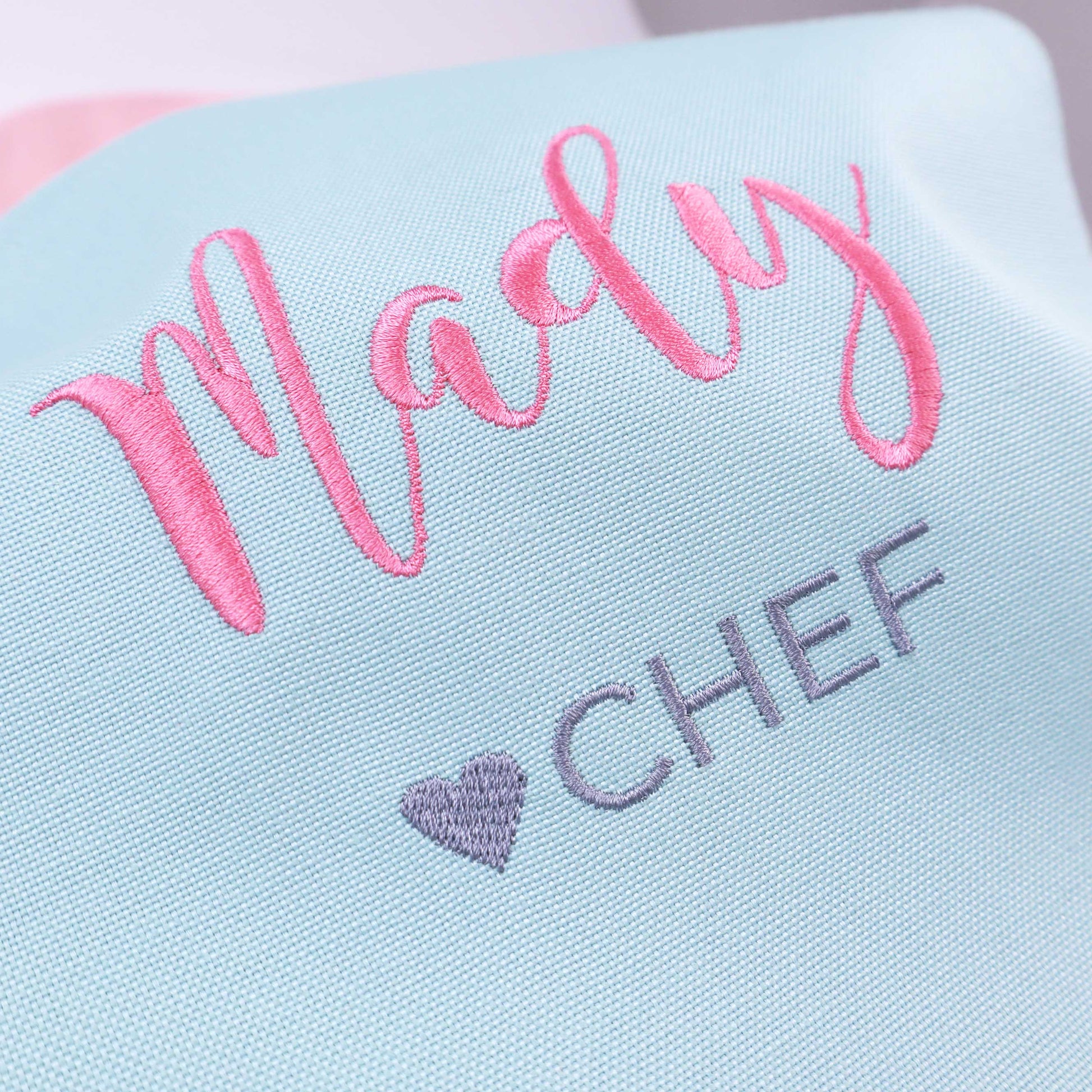 A close-up of a light blue fabric item with pink embroidered script reading 'Madly' and gray embroidered text reading 'CHEF' with a small gray heart.