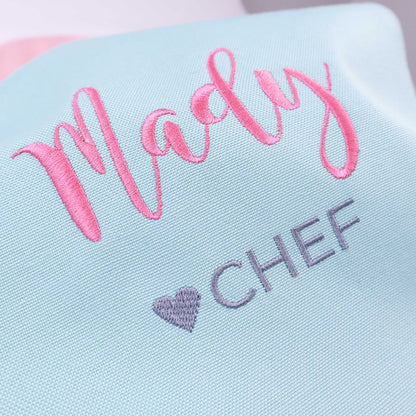 A close-up of a light blue fabric item with pink embroidered script reading 'Madly' and gray embroidered text reading 'CHEF' with a small gray heart.