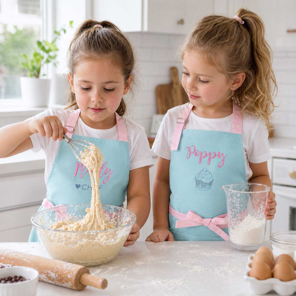 Two young girls in a kitchen wearing matching aprons, one named Poppy, mixing ingredients.