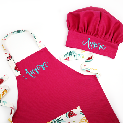 Personalized hot pink apron and chef hat set with a strawberry print, embroidered with the name "Aurora" in teal script