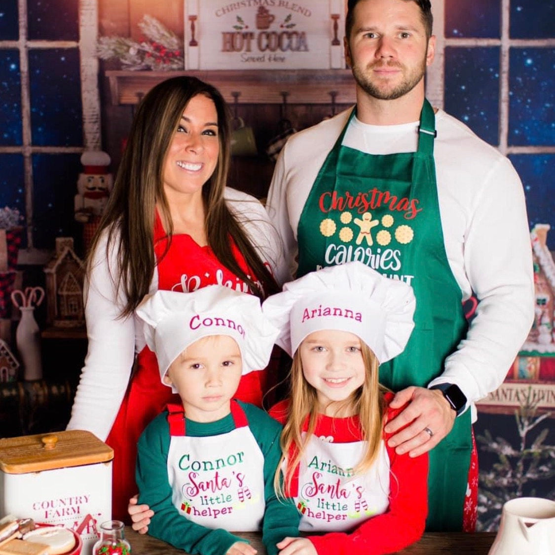 Family of four wearing Christmas-themed aprons and chef hats in a festive kitchen setting.