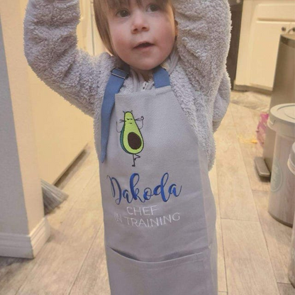 Child wearing a personalized grey apron with an avocado icon, embroidered "Dakota, Chef in Training"