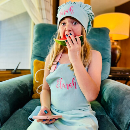 Girl eating watermelon while wearing a personalized mint apron and chef hat, embroidered with the name "Elarah", sitting in an armchair
