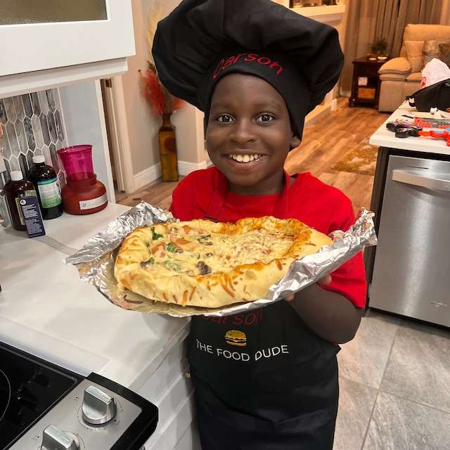 Smiling boy wearing black personalized apron and chef hat holding homemade pizza – The Food Dude design