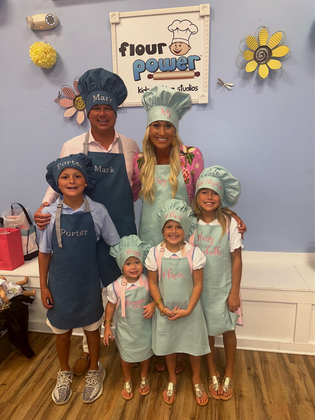 A family of six posing together in matching chef aprons and hats inside  bakery studio. The set includes chef hats and aprons in light blue and navy blue, personalized with names embroidered.