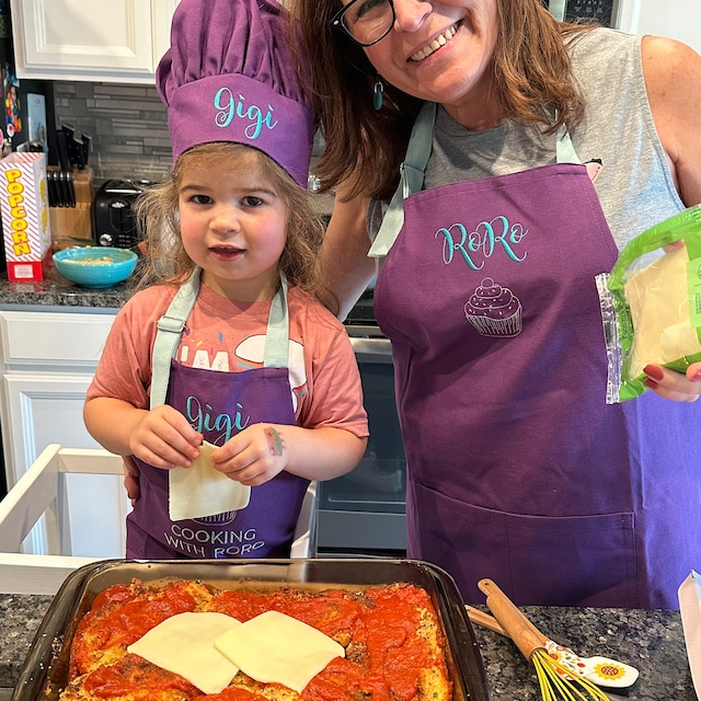 Woman and child in a kitchen wearing matching purple aprons with embroidered names, preparing a dish.