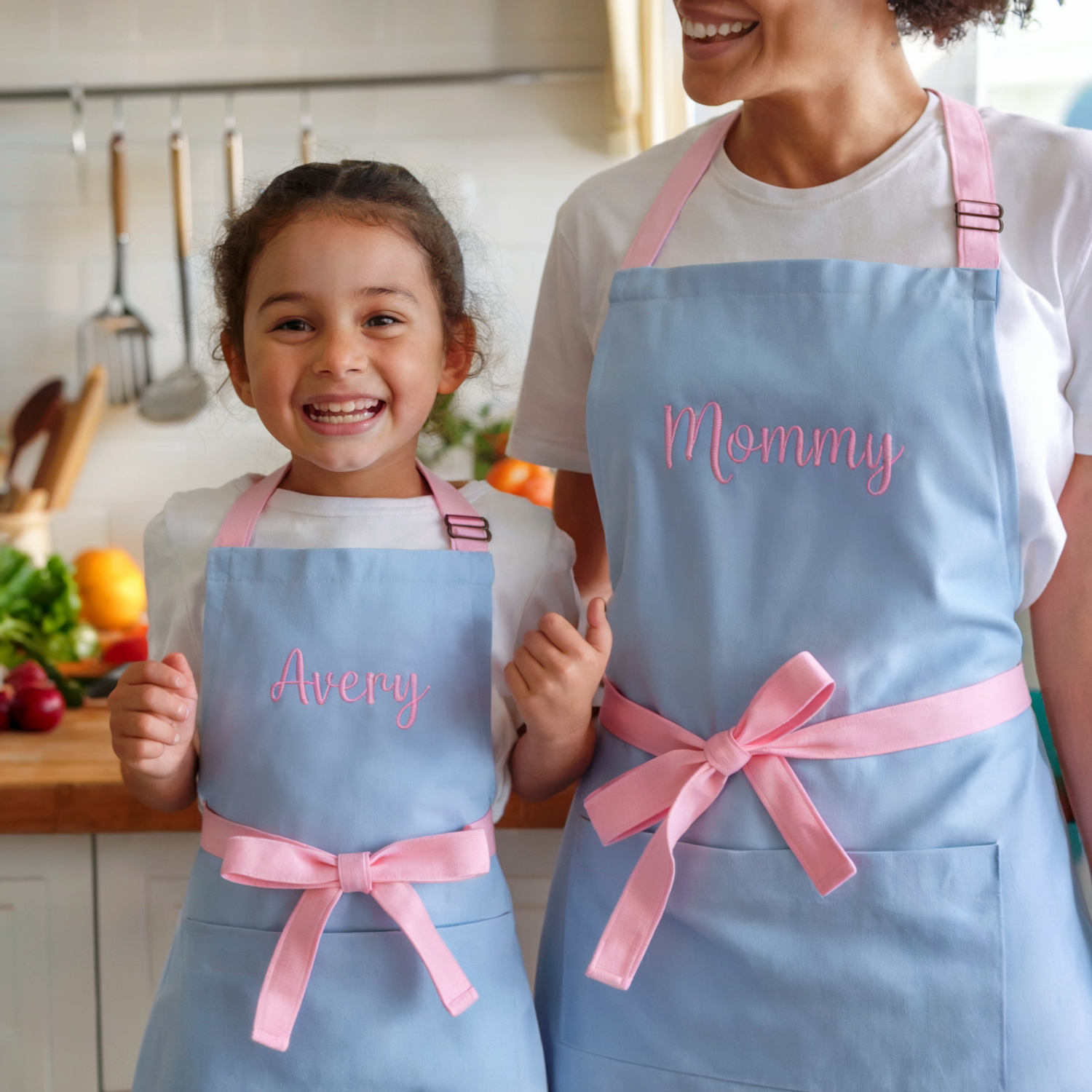 Mother daughter wearing matching blue aprons with pink ribbons and embroidered names in a kitchen.