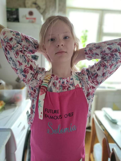 Child reaching up to tie a chef hat behind her head, wearing a floral top