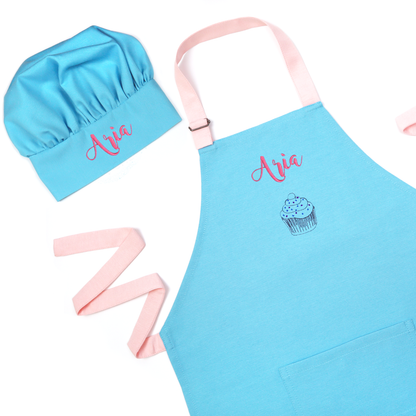 Personalized blue apron and chef hat set with pink straps, embroidered with the name "Aria" above a cupcake icon