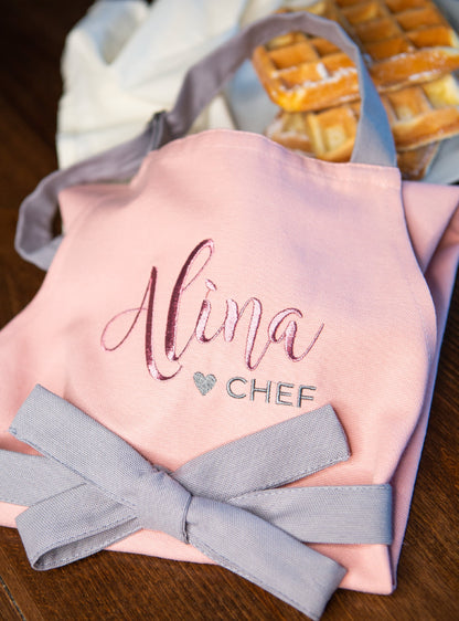 Close-up of a personalized pink apron embroidered with the name "Alina" and a heart "CHEF" design